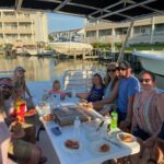 Gathered around a table that is on a boat, getting ready to eat pizza